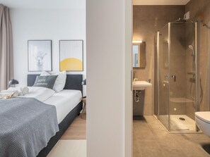 A hotel room with a bed, a bedside table, a lamp, a framed picture, and a shower area with a glass door. - Limehome Frankfurt Gutleutstraãÿe (Frankfurt)