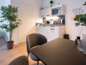 A modern kitchen with white cabinets, a microwave, and a sink. There is a dining table with chairs, a potted plant, and a vase with reeds. - Limehome Frankfurt Gutleutstraãÿe (Frankfurt)