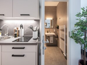 A modern kitchen with a sink, stove, and cabinets, leading to a bathroom with a sink and mirror. - Limehome Frankfurt Gutleutstraãÿe (Frankfurt)
