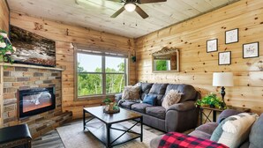 A cozy cabin interior with a stone fireplace, a ceiling fan, a large window, and a comfortable seating area. - Private Indoor Pool/Pet Stays Free/HotTub/Movie (Sevierville)