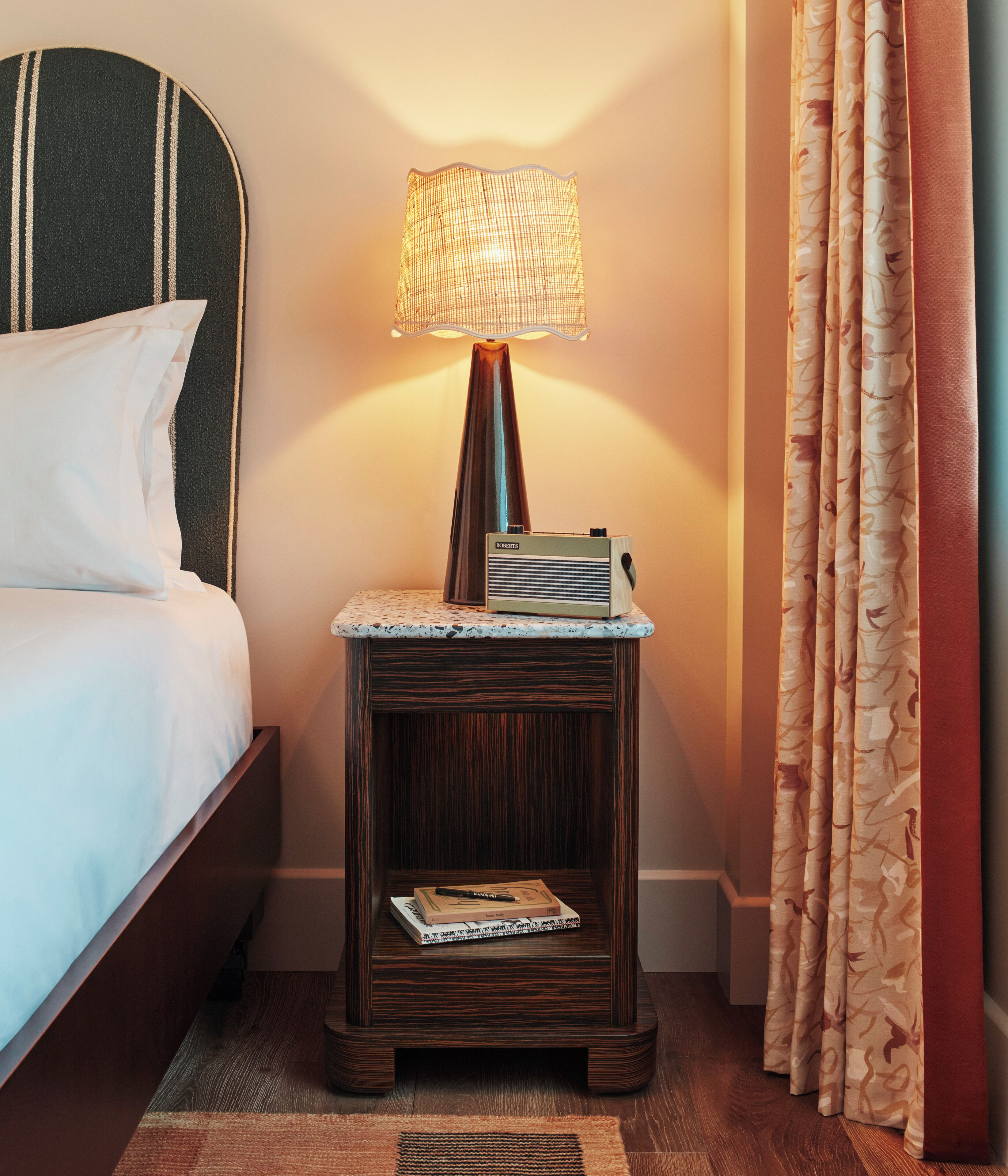 A neatly made bed with pillows, a small bedside table with a lamp, a stack of books, and a curtain.
