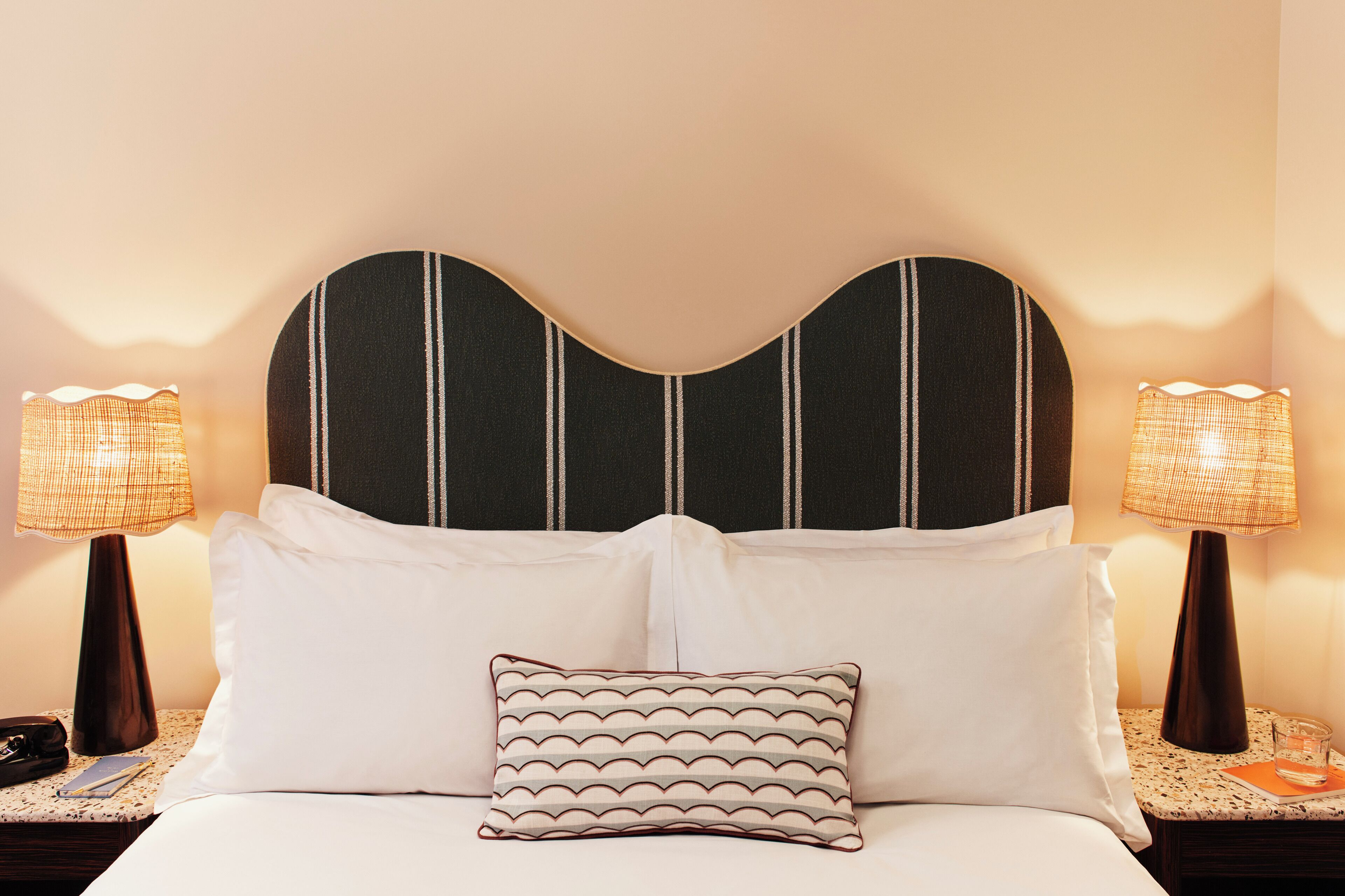 Una habitación de hotel con una cama, dos mesitas de noche, dos lámparas y un cojín decorativo.