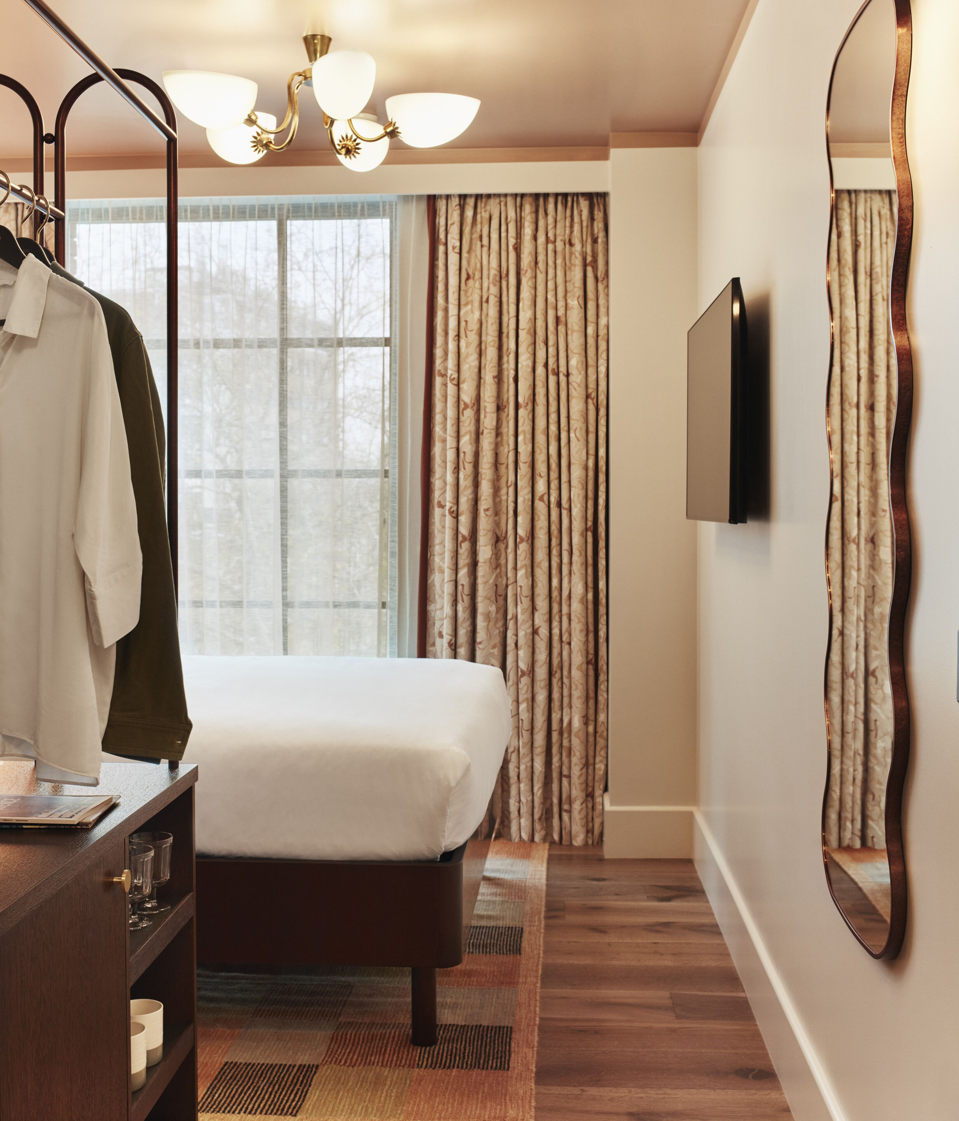 A hotel room with a bed, a wooden dresser, a mirror, a window with curtains, and a wall-mounted TV.