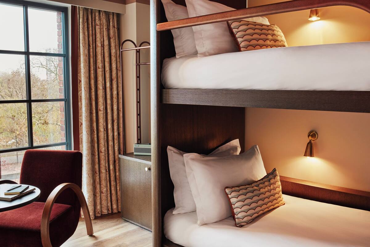 A bunk bed with white bedding and patterned pillows, a wooden desk with a chair, and a window with curtains.