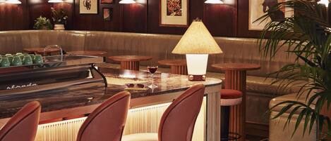 A bar area with a counter, stools, and a lamp. There are bottles on the shelves and a plant in the corner.