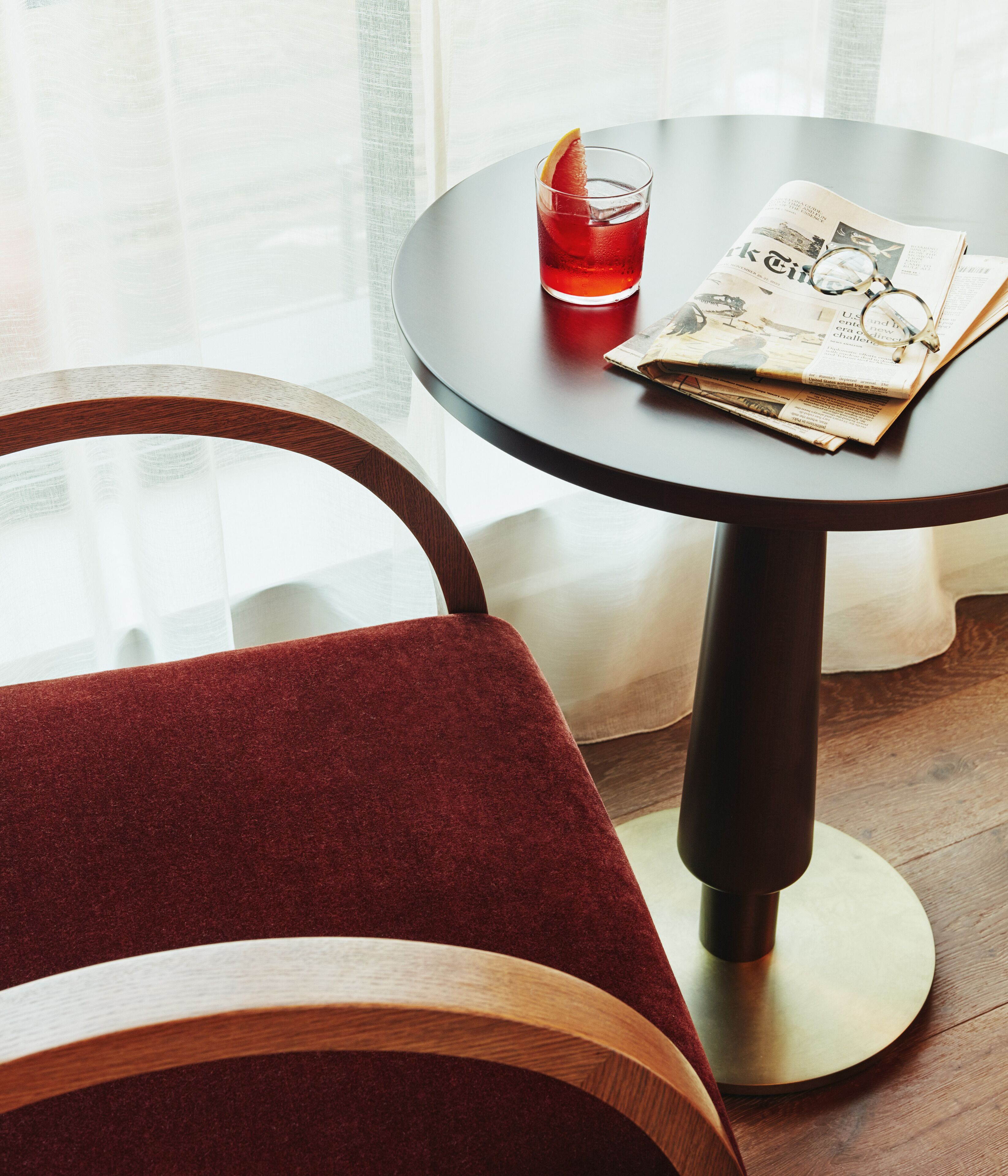 Una mesa redonda con un periódico, un vaso de bebida roja y un par de gafas.