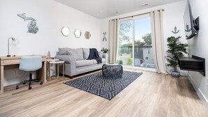 A modern living room with a grey sofa, a small round coffee table, a desk with a chair, and a large window with curtains. - King Bed + Sofa I Walk DT + Front St (Traverse City)