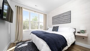 A bedroom with a bed, a TV, a window with curtains, a wall-mounted artwork, and a bedside table with a lamp. - King Bed + Sofa I Walk DT + Front St (Traverse City)