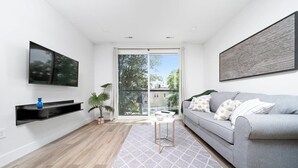 Ein modernes Wohnzimmer mit grauem Sofa, einem Flachbildfernseher, einem Couchtisch und einem großen Fenster mit Blick auf Bäume und Gebäude.