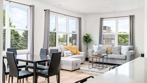 Una sala de estar moderna con comedor, ventanas amplias y un sofá gris con cojines.
