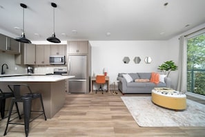 A modern kitchen with a central island, a dining area with a sofa and table, and a large window providing a view of the outdoors.