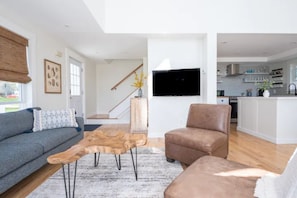 Un salon moderne avec un canapé gris, un fauteuil en cuir marron et une table basse en bois. Un téléviseur à écran plat est fixé au mur, et un escalier mène à un autre niveau.