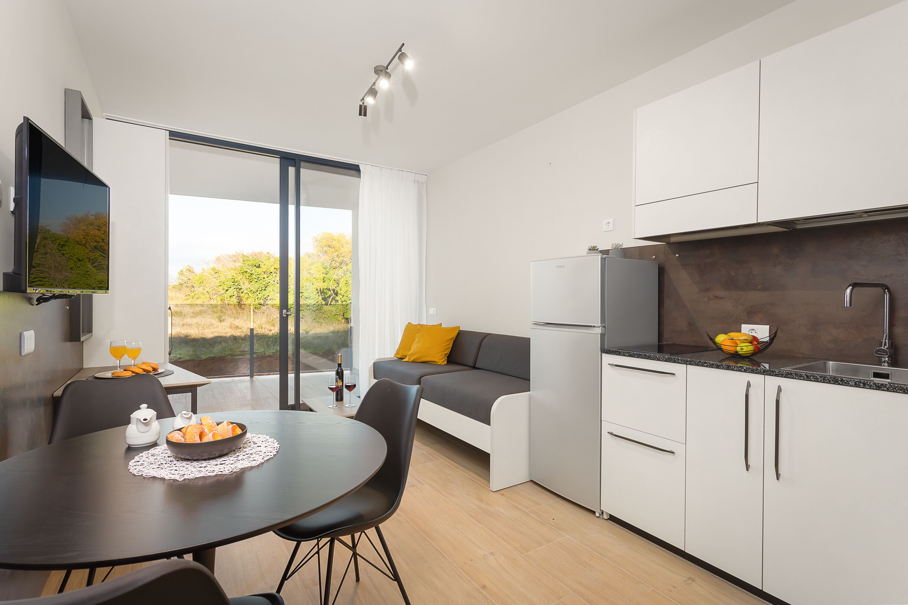 Una cucina moderna con zona pranzo, un divano con cuscino giallo e un'ampia finestra vista alberi.