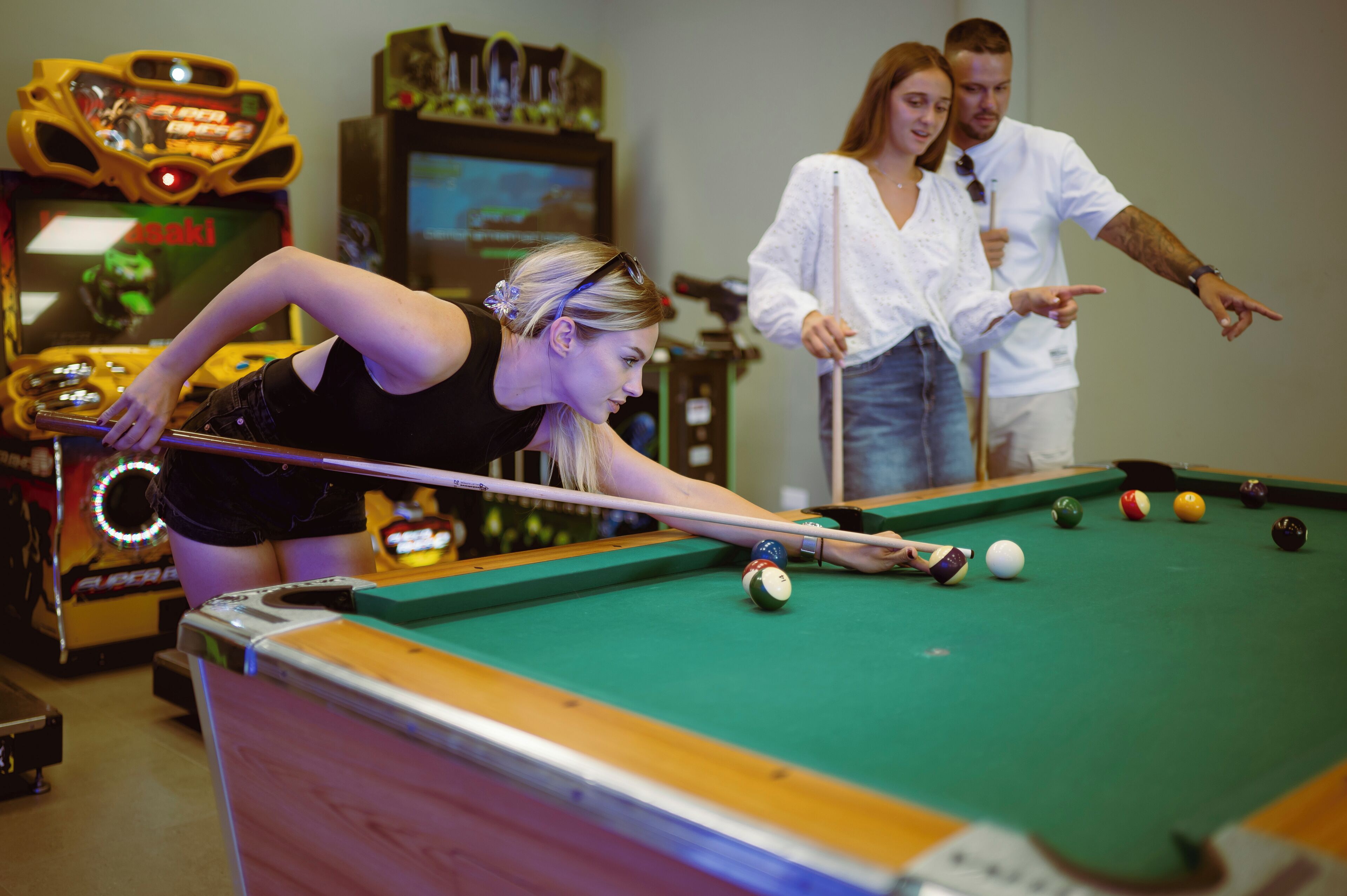 Una mesa de billar con bolas y taco, una persona jugando al billar, y máquinas recreativas al fondo.