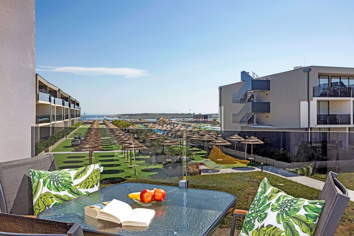 A balcony with a glass table, a book, and fruit. The view includes a pool area with umbrellas and a building with a staircase.