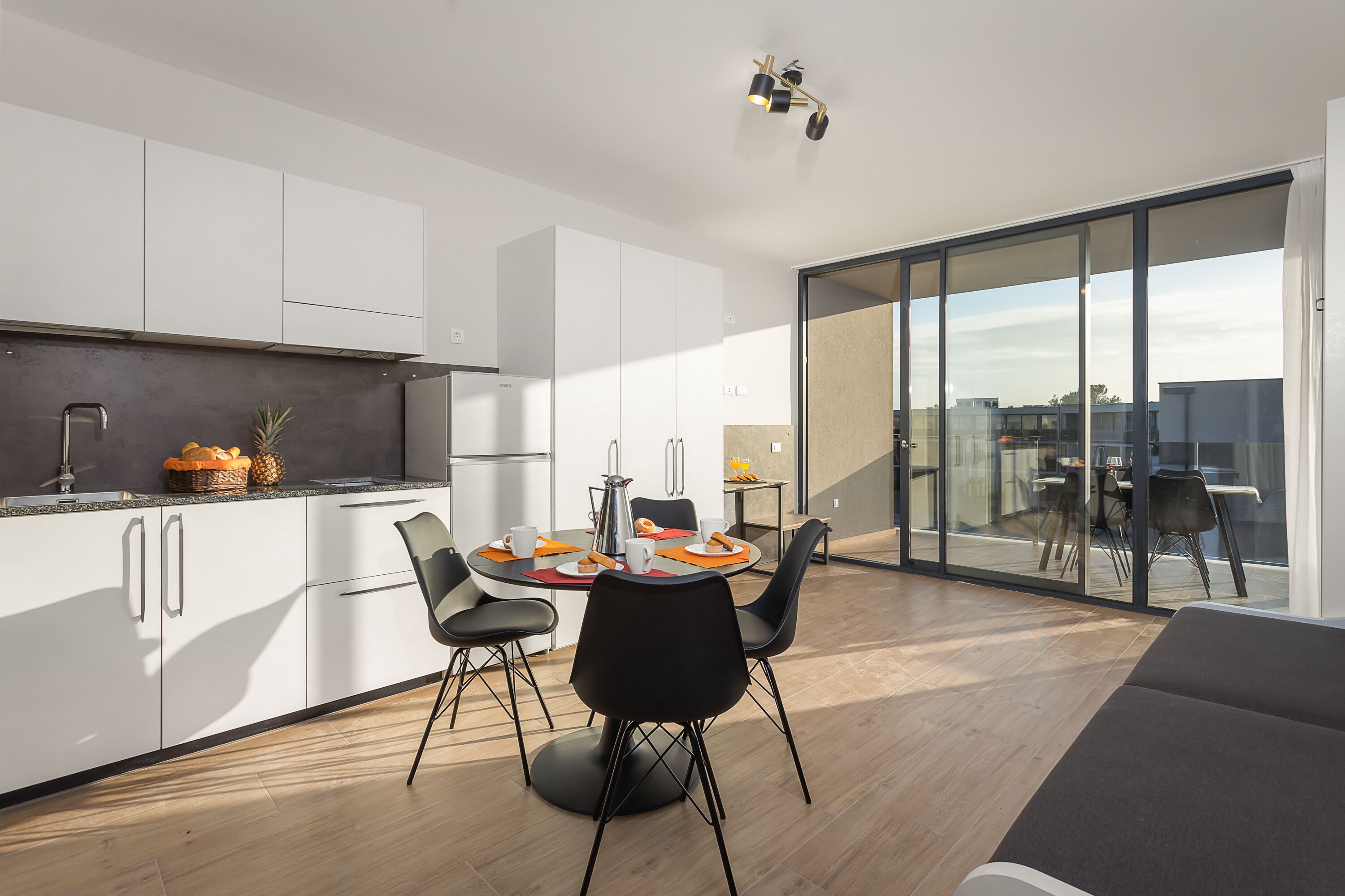 A modern kitchen with white cabinets, a dining area with a round table and black chairs, and a large sliding glass door leading to an outdoor balcony.