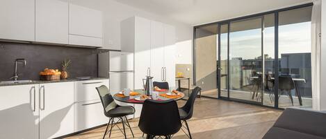 A modern kitchen with white cabinets, a dining area with a round table and black chairs, and a large sliding glass door leading to an outdoor balcony.