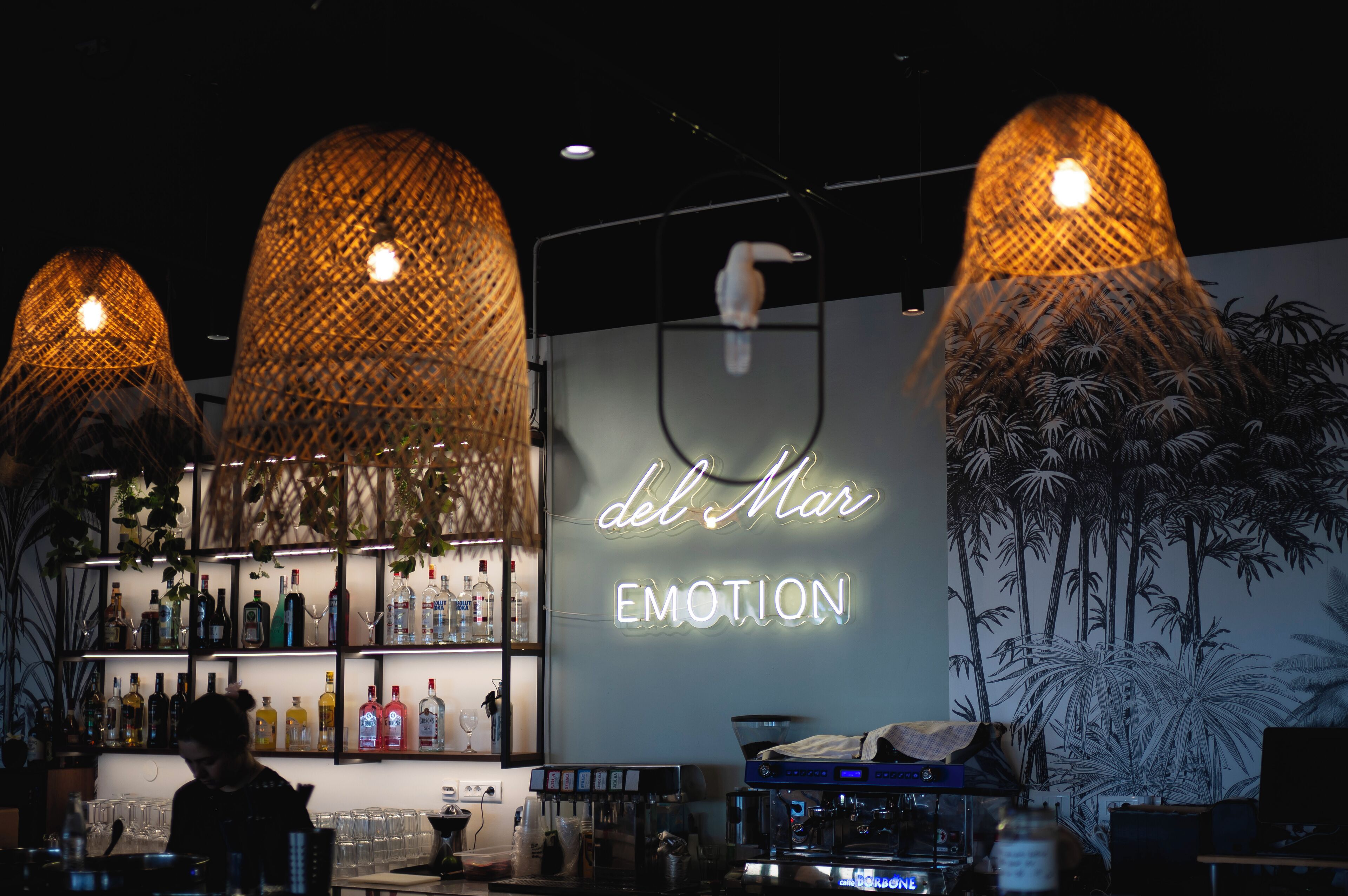 A bar area with a neon sign reading "del Mar EMOTION," shelves with bottles, and a mural of palm trees.