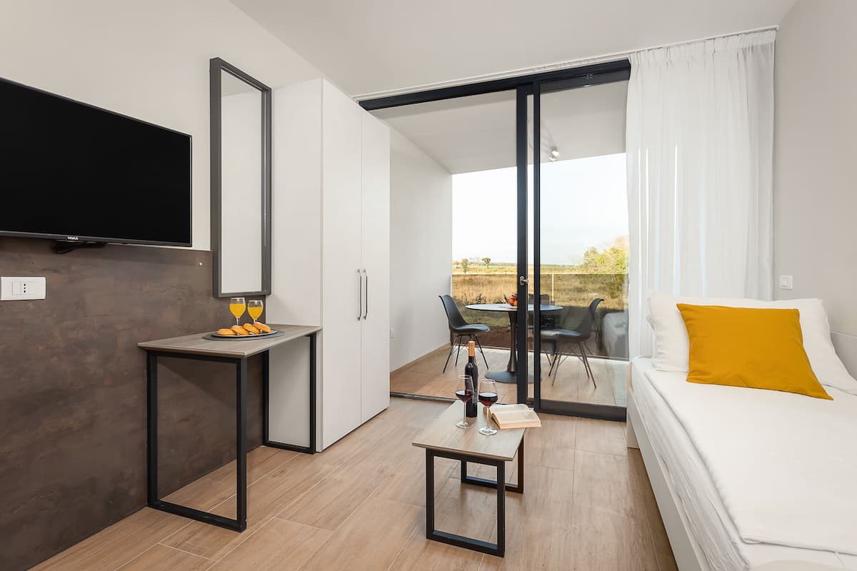 A modern hotel room with a flat-screen TV, a small table with a bottle of wine and glasses, a sofa with a yellow pillow, and a sliding glass door leading to a balcony with a table and chairs.
