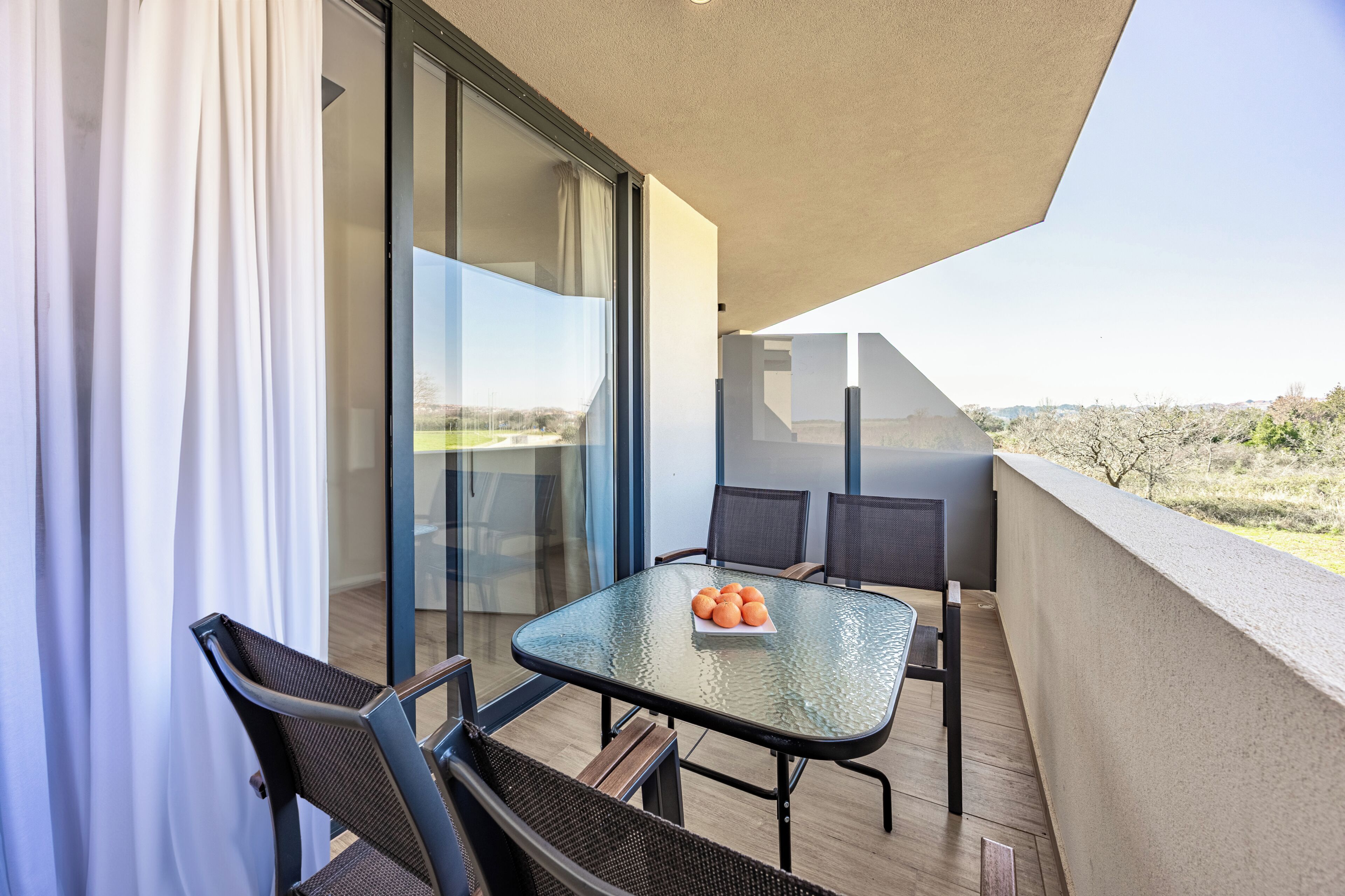 A balcony with a glass table, chairs, and a view of the landscape.