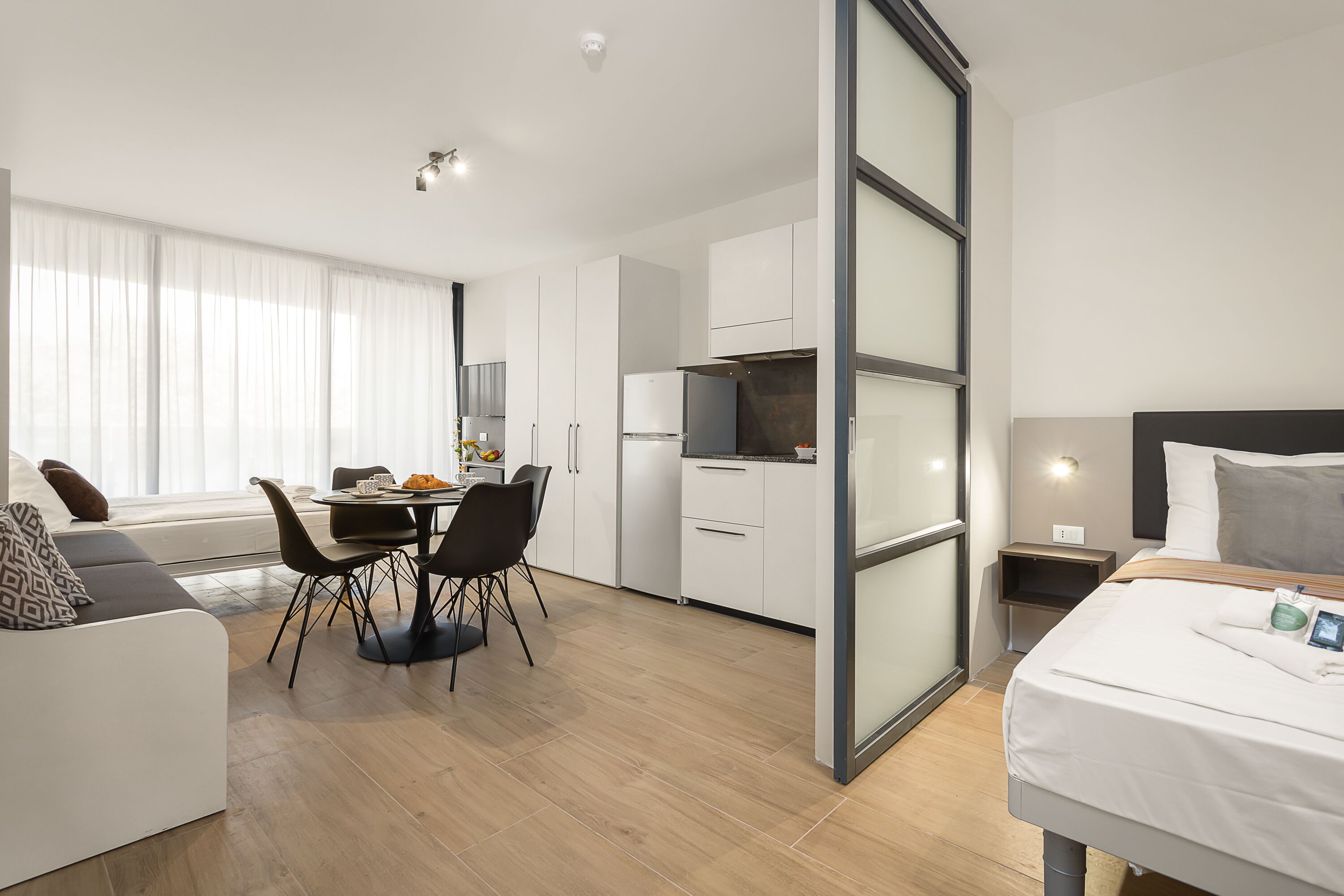 A modern hotel room with a bed, a dining area with a table and chairs, a kitchen area with a refrigerator and stove, and a large window with curtains.