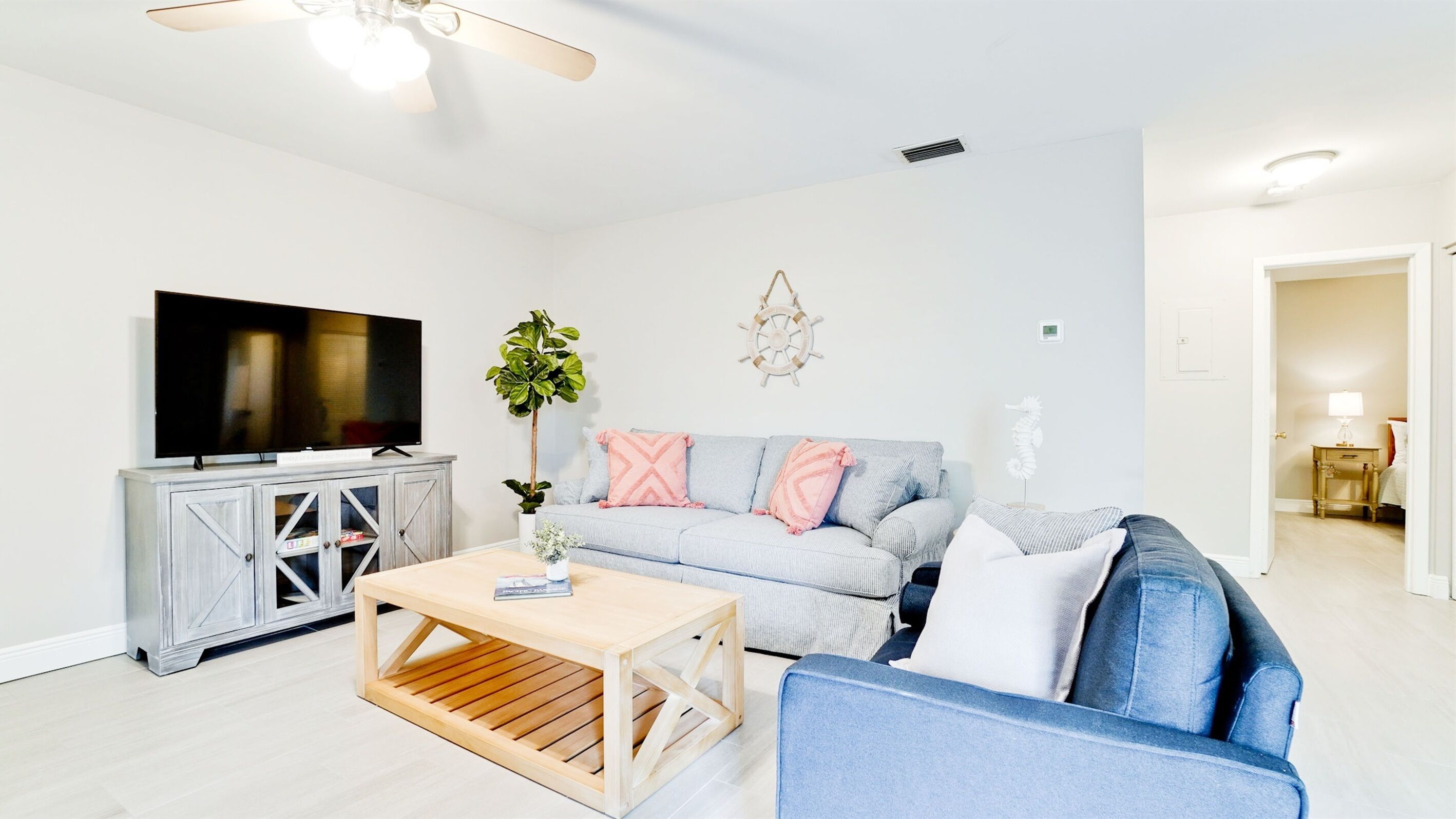 A modern living room with a flat-screen TV, a wooden coffee table, a grey sofa with patterned cushions, and a potted plant.