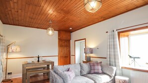 Una sala de estar acogedora con techo de madera, un sofá gris, un banco de madera, una mesa con una lámpara y una ventana con cortinas.