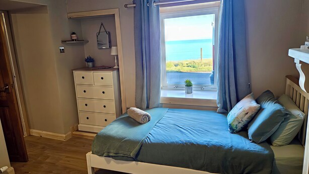 Un dormitorio con cama, cómoda, ventana con vista al mar y una pequeña estantería con una planta.