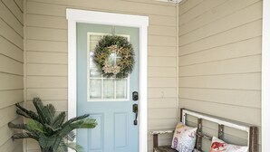 Uma porta azul clara com uma coroa de flores, uma planta em vaso e um banco com almofadas.