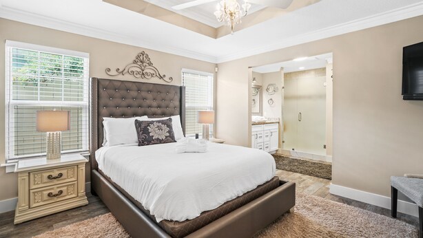 Quarto com cama grande, um lustre, televisão e banheiro visível através de uma porta aberta.