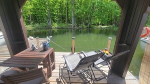 A wooden deck with a table and chairs, overlooking a calm body of water and dense greenery. - Aloberge 1 (Lacolle)