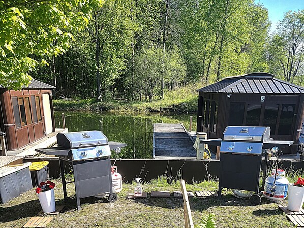 Una cabaña junto al lago con parrillas exteriores, un cilindro de propano y un cobertizo pequeño.