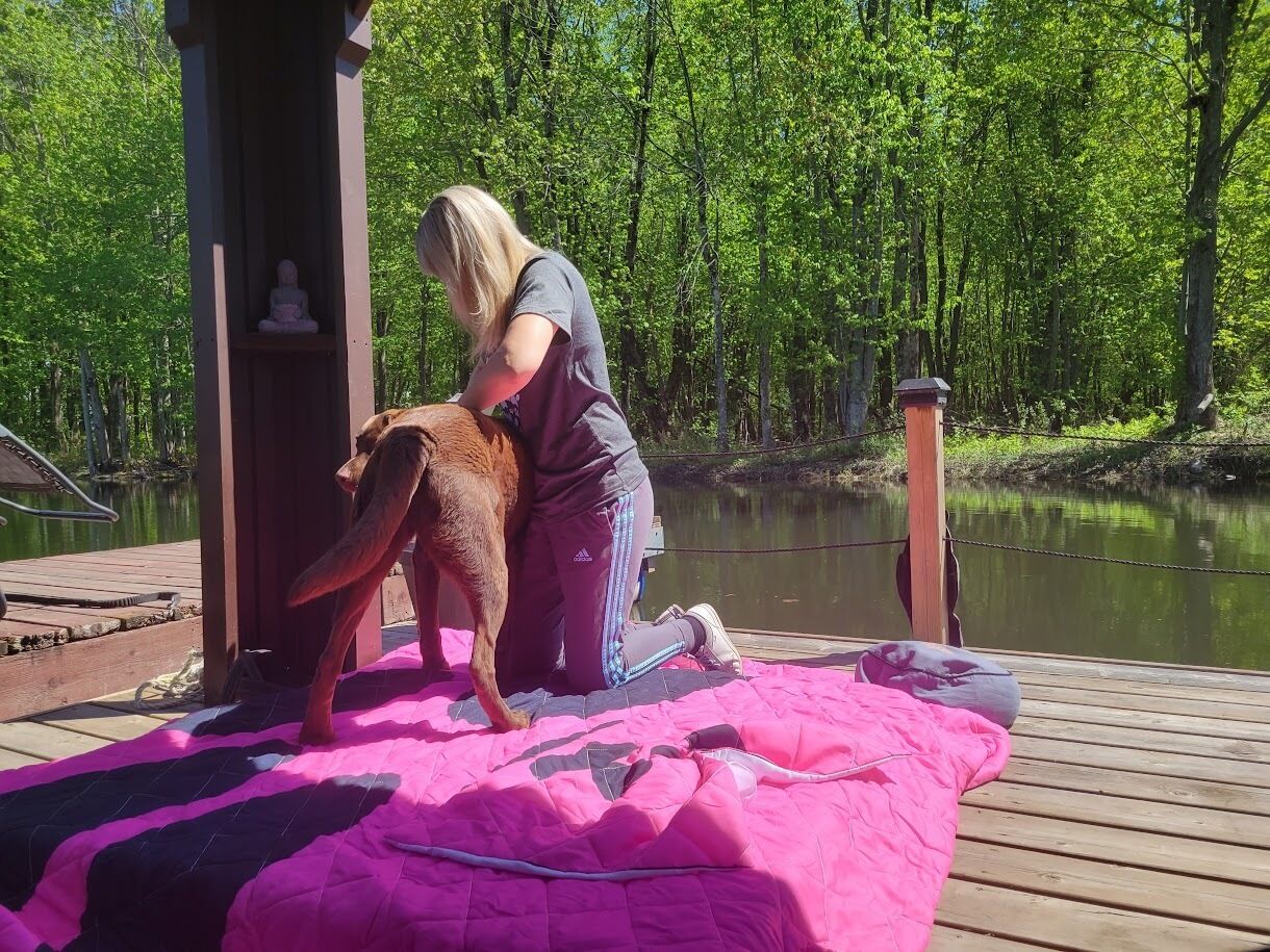 A person on a wooden deck with a dog, a pink blanket, and a small statue.
