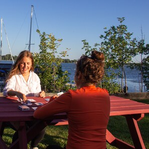 Two people playing cards at an outdoor table with a view of a body of water and boats in the background.