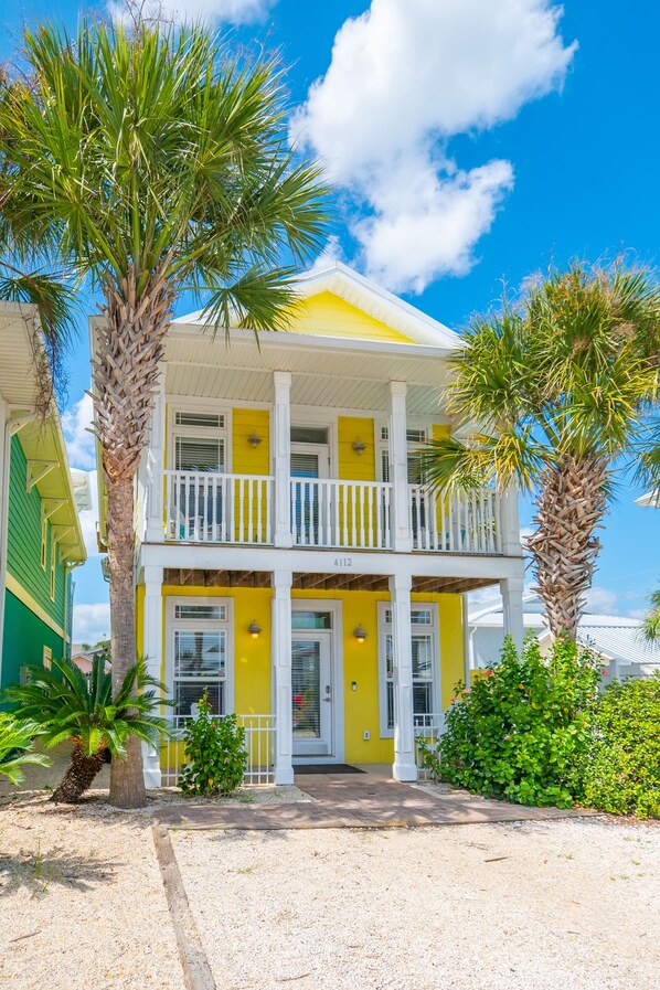 A two-story yellow building with a balcony, surrounded by palm trees and a gravel driveway.