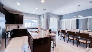 A modern kitchen with dark cabinets, a marble countertop, and a dining area with a large window. - Beautiful home across from the Clubhouse! 5 Star Reviews! Fun Resort!  (Kissimmee)