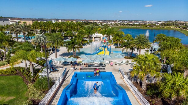 A water park with a large pool, palm trees, and a view of a lake and houses in the background. - Beautiful home across from the Clubhouse! 5 Star Reviews! Fun Resort!  (Kissimmee)