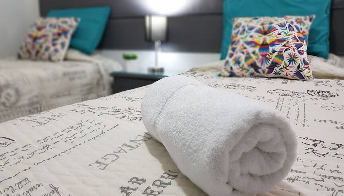Ein ordentlich bezogenes Bett mit einem gerollten weißen Handtuch, einem türkisfarbenen Kopfkissen und einem gemusterten Kissen auf einem Bett mit einem bedruckten Bettlaken.