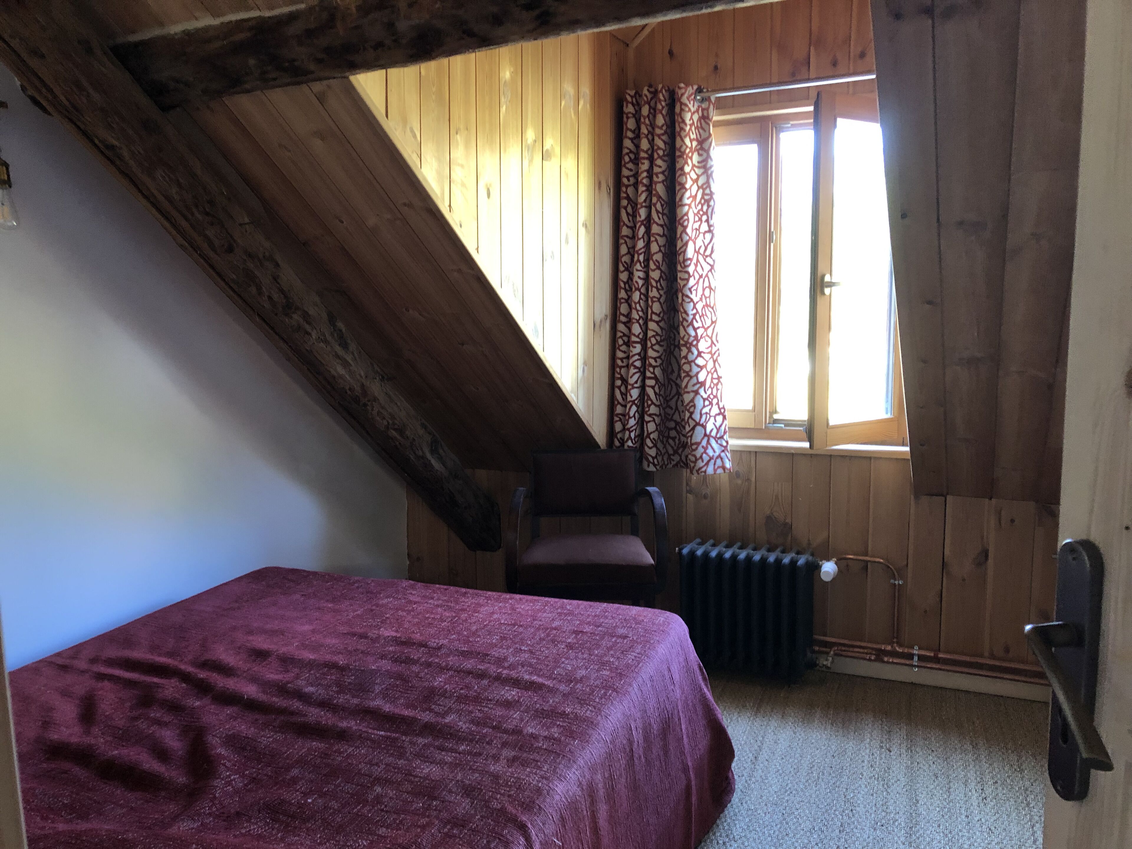 A room with a sloped ceiling, a bed with a red bedspread, a chair, a radiator, and a window with curtains.