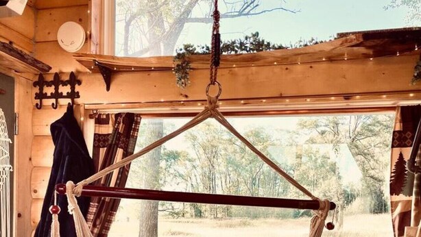 A hammock suspended from a wooden ceiling in a rustic cabin with a view of a grassy landscape.