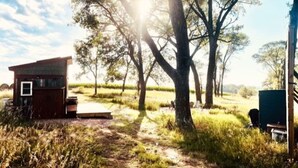 A rustic cabin with a small porch and a pathway leading to it, surrounded by trees and open fields.