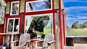 A red wooden cabin with two windows and a door, featuring a small table and two chairs outside.