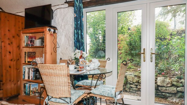 A cozy room with a glass table, wicker chairs, a bookshelf, and a view of a garden through a sliding glass door. - Carminow Barn (Bodmin)