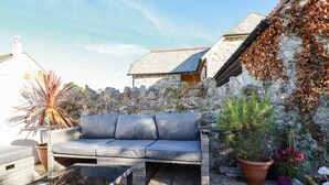 A patio area with a grey sofa, a glass-top table, and potted plants. - Carminow Barn (Bodmin)