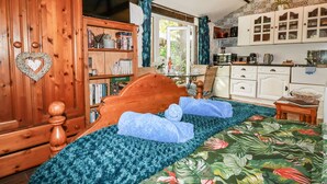 A bedroom with a wooden headboard, a bed with a green floral bedspread, a bookshelf, and a kitchen area with white cabinets and a microwave. - Carminow Barn (Bodmin)