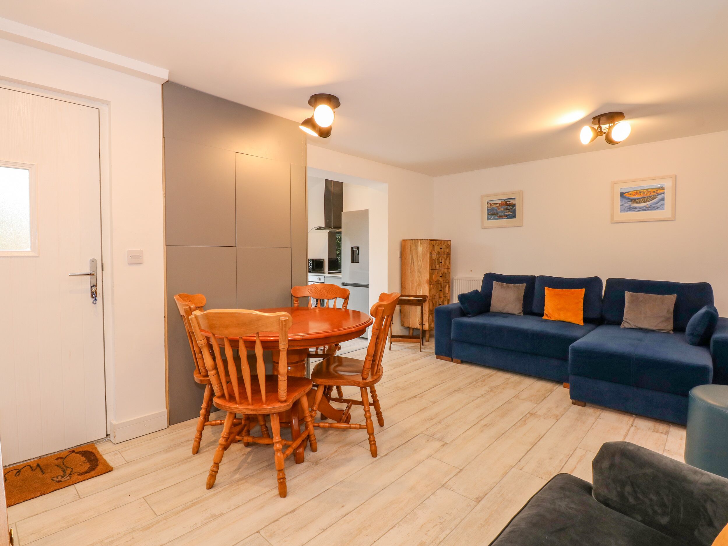 Een moderne woonkamer met een blauwe bank, een houten eettafel en stoelen. Achtergrond toont een keuken met een koelkast en kasten.