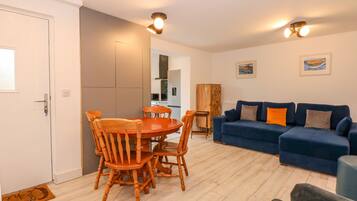 Een moderne woonkamer met een blauwe bank, een houten eettafel en stoelen. Achtergrond toont een keuken met een koelkast en kasten.