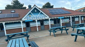 A beach cafe with a blue and white exterior, solar panels on the roof, and multiple picnic tables in the foreground.