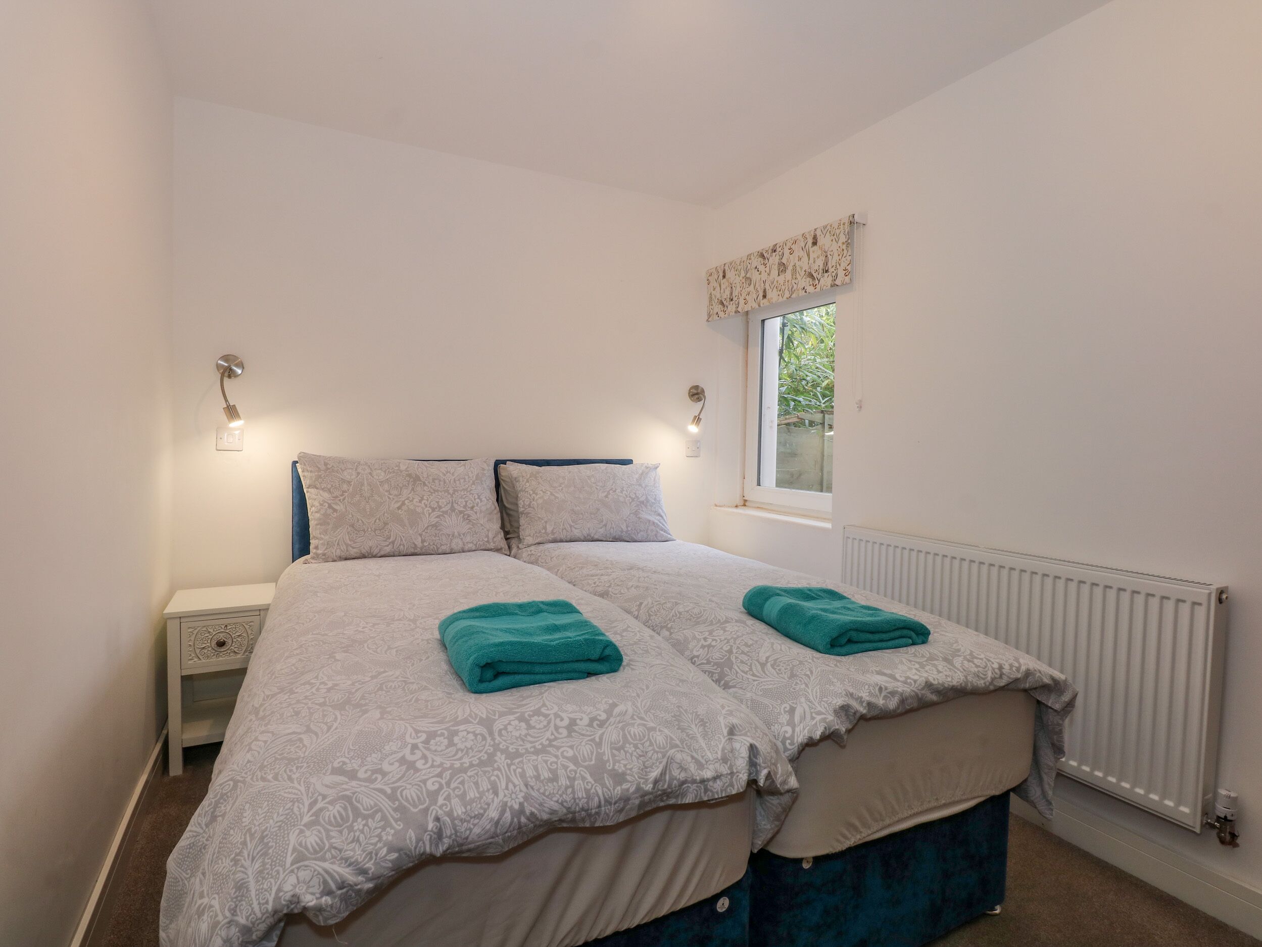 A double bed with patterned bedding and two teal towels, a bedside table, a nightstand, a window with a view, and a wall-mounted light fixture.