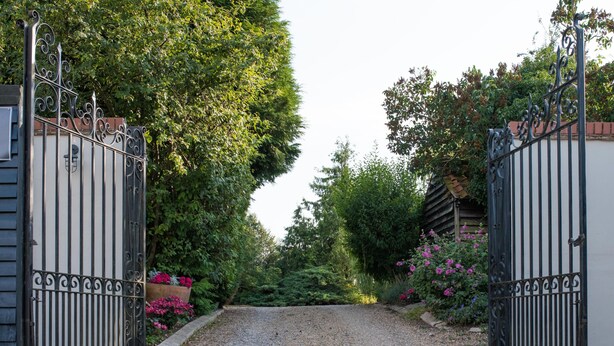 Une allée de gravier mène à une entrée fermée par un portail en métal, avec un bâtiment en bois en arrière-plan.